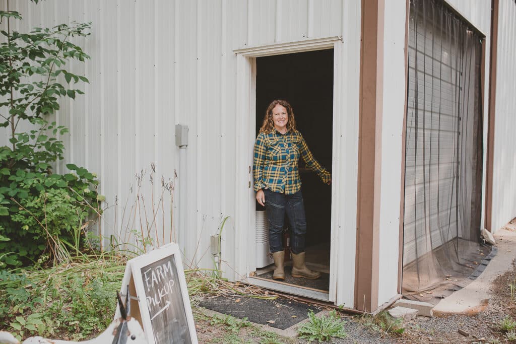 Leslie standing in the doorway of her farm store, smiling at the camera and wearing jeans, rubber boots and a plaid shirt
